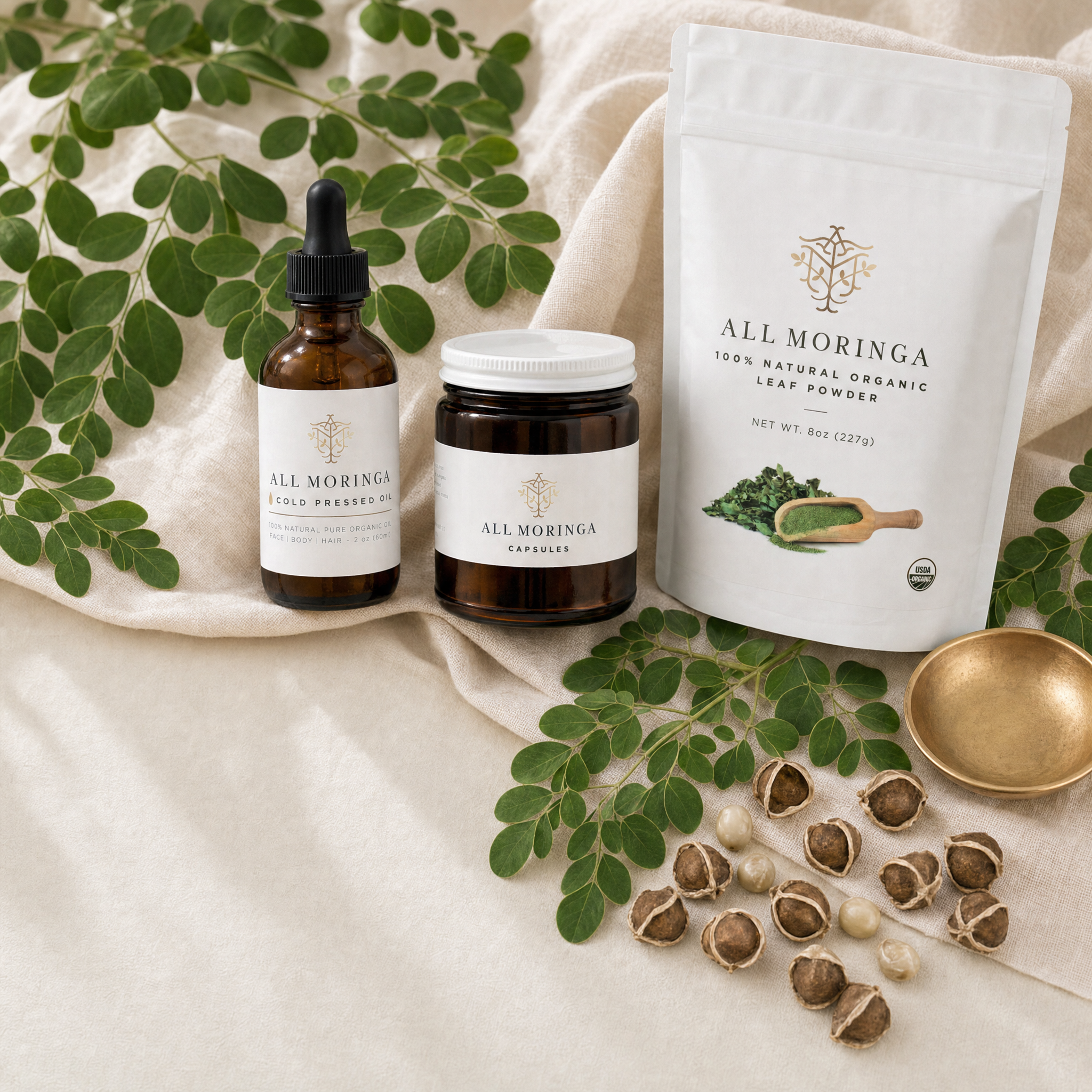 All Moringa starter kit on warm linen: cold-pressed seed oil bottle, capsules jar and leaf powder pouch with fresh moringa leaves and seeds