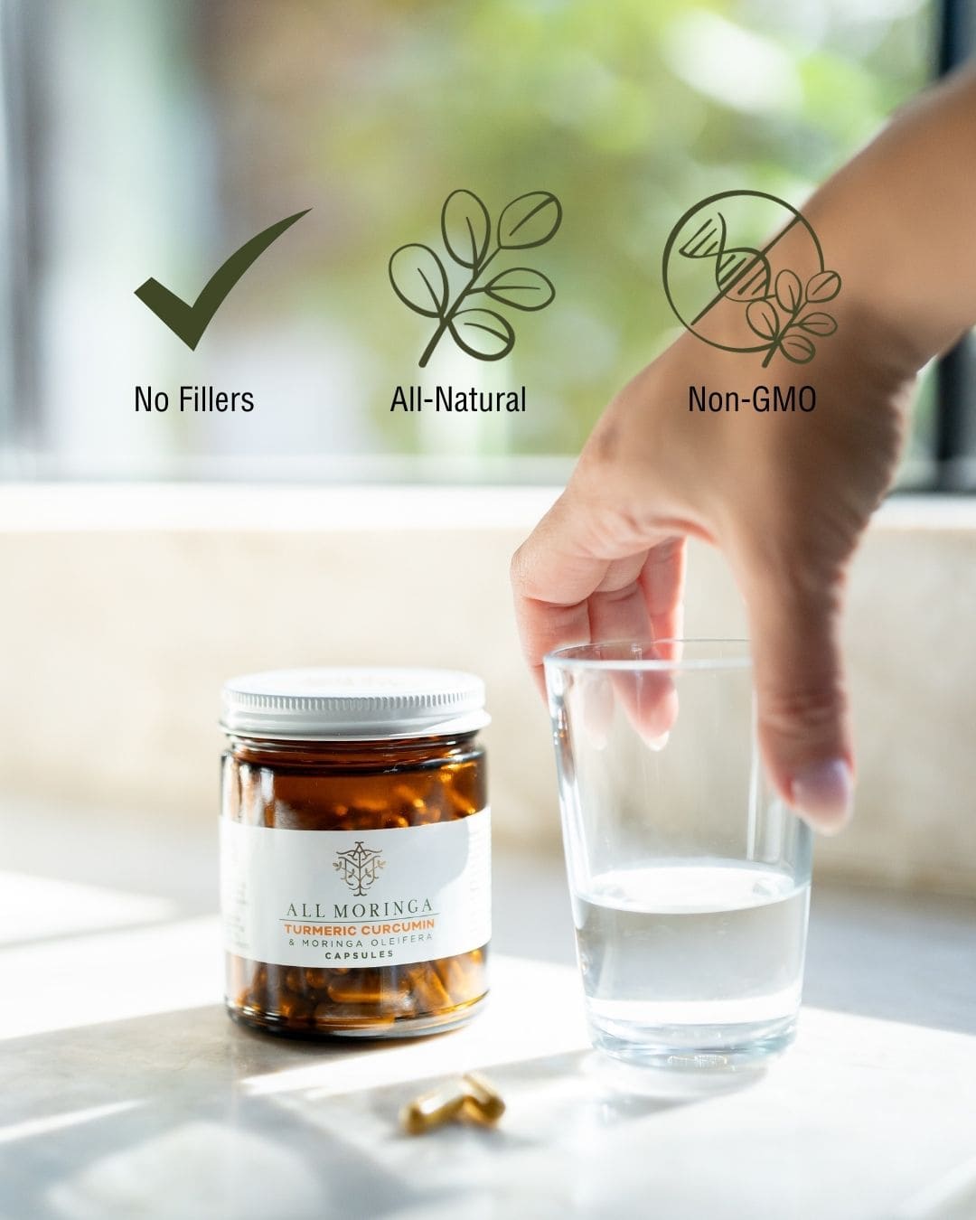 Jar of turmeric moringa capsules, glass of water, and hand reaching for a capsule with 'No Fillers', 'All-Natural', and 'Non-GMO' icons.